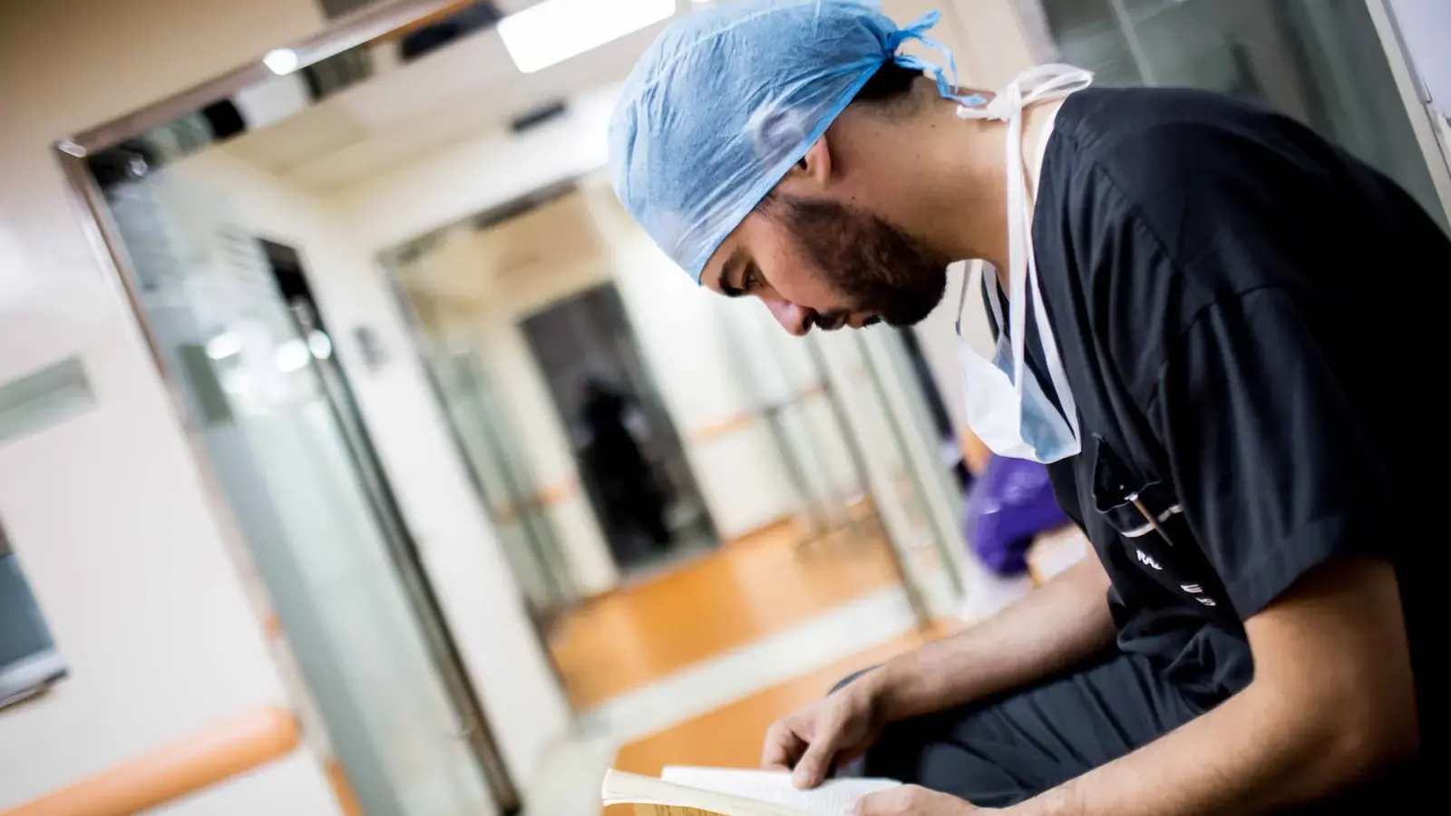 A doctor in scrubs and a surgical cap reading a book in a hospital corridor