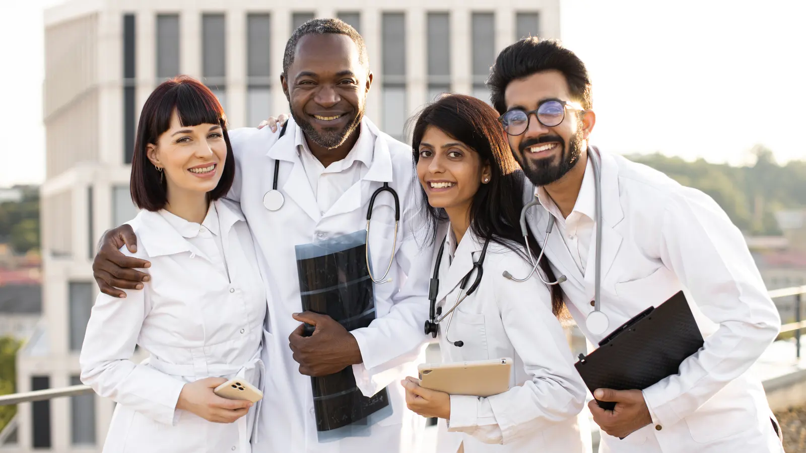 Four smiling doctors in white coats standing together outside a hospital