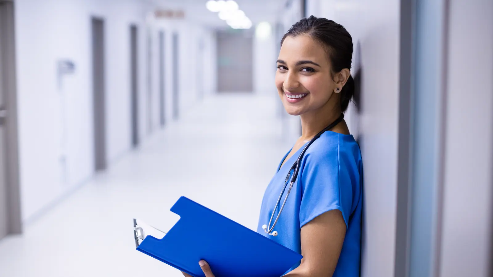 A medical student in scrubs holding a patient file in a hospital corridor