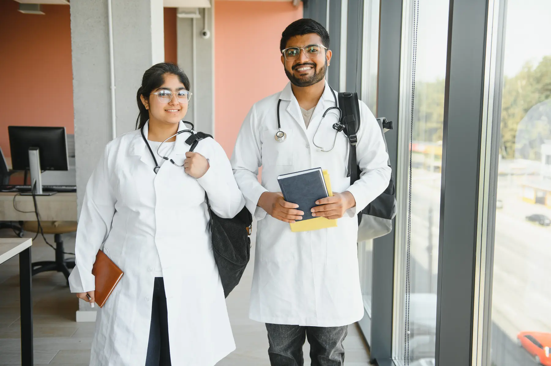 Two medical students in white coats with stethoscopes and backpacks, smiling in a hospital corridor